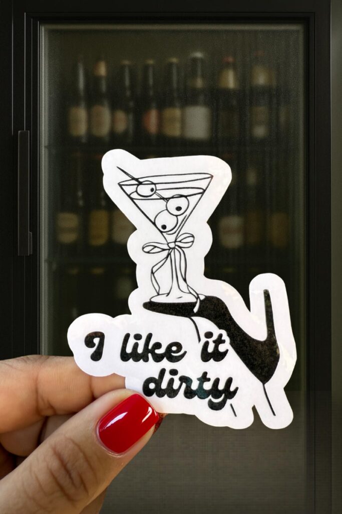 Black-and-white sticker of a martini glass with three olives tied in a ribbon, balanced on a high heel, captioned “I like it dirty.”
