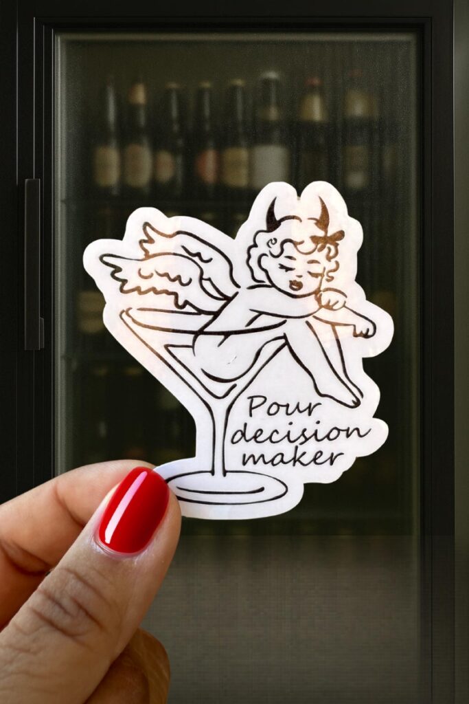 Cartoon sticker of a cherub lounging inside a martini glass, with the pun “Pour decision maker.”